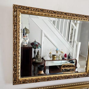 May include: A rectangular wall mirror featuring a wide, ornate gold-colored frame with intricate embossed scrollwork and floral detailing. The reflective glass surface is clear and smooth, displaying a reflection of an interior room setting.