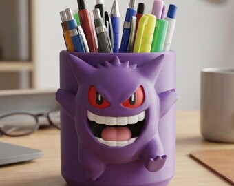May include: A purple cylindrical pencil holder featuring a molded 3D Gengar character with red eyes, a wide white-toothed grin, and a pink tongue. The container is filled with various colorful pens and markers, including blue, red, and yellow.