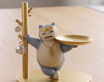 May include: A gold-colored desk organizer featuring a cartoon character wearing sunglasses and holding a tray. The organizer has a tree-like structure for hanging jewelry and a round base. Two pieces of jewelry are hanging from the tree.