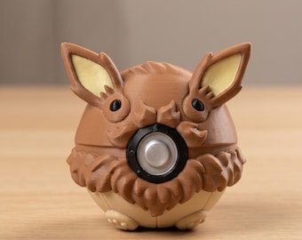 May include: A brown and tan spherical figurine with large ears and a furry collar. The figurine has a black circular feature in the center and is set on a wooden surface. The ears are tan with a yellow interior.