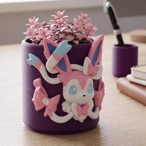 Puede incluir: Una maceta cilíndrica morada con un diseño de Sylveon, que contiene plantas rosas. La figura de Sylveon es blanca, rosa y azul. La maceta está sobre un escritorio de madera con un portalápices y cuadernos.