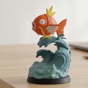 Puede incluir: Figura detallada de un pez de dibujos animados naranja con detalles amarillos y blancos, sobre olas azules estilizadas. El pez tiene una expresión de sorpresa. La base es negra y la figura está sobre una superficie clara.