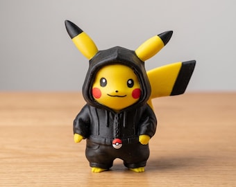 May include: A yellow Pikachu figurine wearing a black hooded jacket and pants. The figurine has red cheeks, black-tipped ears, and a tail. A red and white Poké Ball is on the jacket. The figure is standing on a wooden surface.
