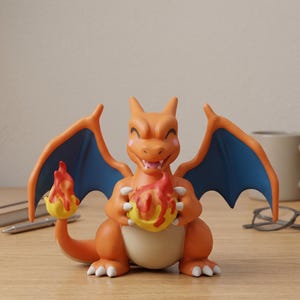 May include: Orange Charizard figurine with blue wings, holding two flames. The figurine has a smiling expression and is set on a wooden surface. A mug, glasses, pen, and notebook are in the background.