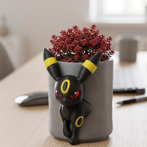 May include: A gray cylindrical planter featuring a black Umbreon figure with yellow accents and red eyes. The planter holds a vibrant red plant. The design is a unique blend of pop culture and home decor.