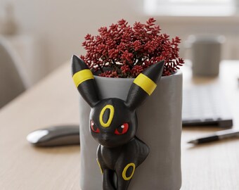 May include: A gray cylindrical planter featuring a black Umbreon figure with yellow accents and red eyes. The planter holds a vibrant red plant. The design is a unique blend of pop culture and home decor.