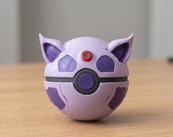 May include: A lavender and purple spherical collectible, resembling a character's head, with a red button on top. The object has a circular design in the center and is resting on a wooden surface.