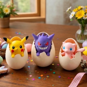 May include: Three Pokémon figures are displayed emerging from eggshell-shaped containers. A yellow Pikachu, a purple Gengar, and a pink Mew are featured. The scene is set on a wooden table with Easter eggs and flowers in the background.