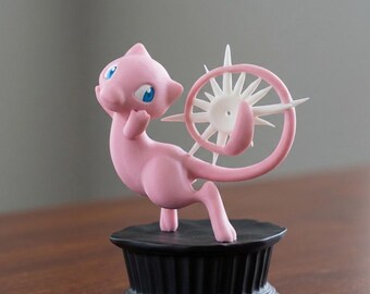 May include: A pink figurine of a cartoon creature with blue eyes and a long tail, holding a white and pink circular design. The figurine is mounted on a black, fluted base. The background is a blurred gray.
