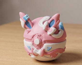 May include: A pink and white decorative sphere with blue accents. The sphere has a detailed design with bow-like features and a circular center. The object is sitting on a wooden surface.