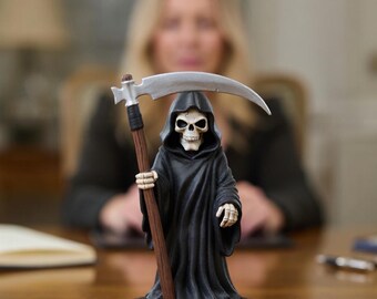 May include: A small figurine of the Grim Reaper, featuring a skeletal face and a black hooded robe. The figure holds a scythe with a silver blade and a wooden handle. The figurine is on a brown base.