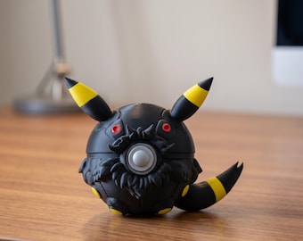 May include: A black and yellow spherical figurine with pointed ears and a tail. The figurine has red eyes and a central silver button. The ears and tail have yellow accents. The object is on a wooden surface.