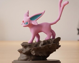 May include: A pink figurine of a cat-like creature with large ears and a long tail, standing on a brown rock formation. The creature has blue accents on its ears and eyes. The figurine is on a brown circular base.