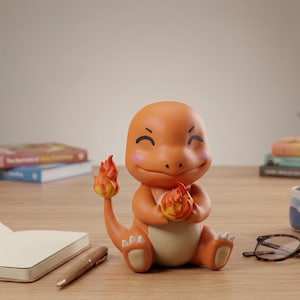 May include: Orange figurine of a Charmander character with a happy expression, holding a flame in its hands. The figurine has a flame on its tail and is sitting on a wooden surface. Other items include a notebook, pen, glasses, and books.