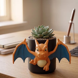 Può includere: Un vaso di ceramica nero con una figura di Charizard attaccata, contenente una pianta succulenta. Il Charizard è arancione con ali blu. Il vaso è su una scrivania di legno.