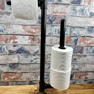 May include: A black metal toilet paper holder with a red valve on top. The holder has two rolls of toilet paper and a wooden base.