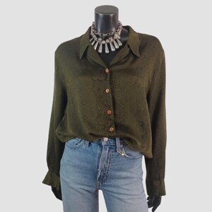 Può includere: Una camicia a maniche lunghe verde oliva con una sottile trama simile alla pelle di serpente, dotata di colletto classico e bottoni chiari. La camicia è abbinata a jeans in denim azzurro e a una vistosa collana color argento.