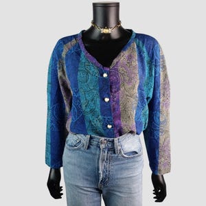 May include: A vintage paisley print blouse in shades of blue, purple, and teal, featuring long sleeves and a V-neck. The blouse, fastened with three pearl-like buttons, is displayed on a black mannequin. It is paired with light wash denim jeans.