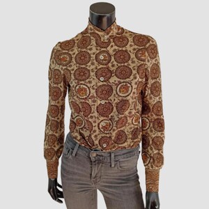 Può includere: Camicia abbottonata beige con un motivo ripetuto di disegni circolari marroni. La camicia a maniche lunghe ha un colletto alto e polsini abbottonati. La camicia è esposta su un manichino.
