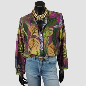 May include: A long-sleeved button-down shirt featuring a vibrant botanical print with large purple, green, and gold leaves and flowers on a dark purple background. The shirt is paired with light blue denim jeans and a gold-toned heart-buckle belt.