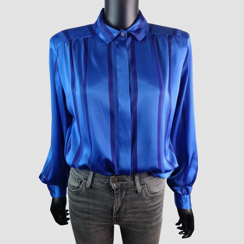 May include: A blue satin blouse with vertical stripes in varying shades of blue. The long-sleeved shirt has a collared neckline and buttoned cuffs. The blouse is displayed on a mannequin.
