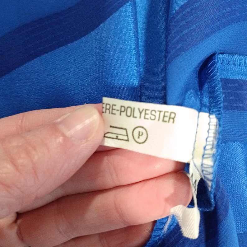 May include: Close-up of a blue fabric with a label that reads "ERE-POLYESTER". The label also features a symbol of an iron and a circle with a "P" inside. The fabric has a striped pattern.