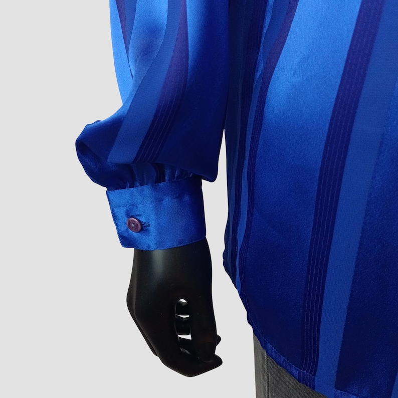 May include: A blue long-sleeve shirt with vertical stripes in varying shades of blue. The cuff features a button closure. The shirt is made of a shiny, silky material.