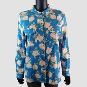 May include: A blue button-down shirt with a floral pattern. The shirt features a repeating pattern of light pink roses and green leaves. It has two chest pockets and long sleeves. The shirt is displayed on a black mannequin.