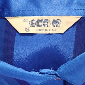 May include: Close-up of a blue collared shirt with a label that reads "44 ECAMB MADE IN ITALY". The shirt has vertical stripes and a buttoned collar.