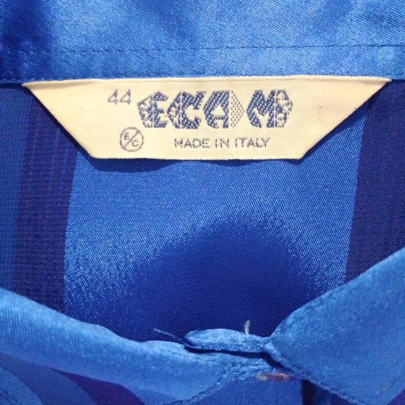 May include: Close-up of a blue collared shirt with a label that reads "44 ECAMB MADE IN ITALY". The shirt has vertical stripes and a buttoned collar.