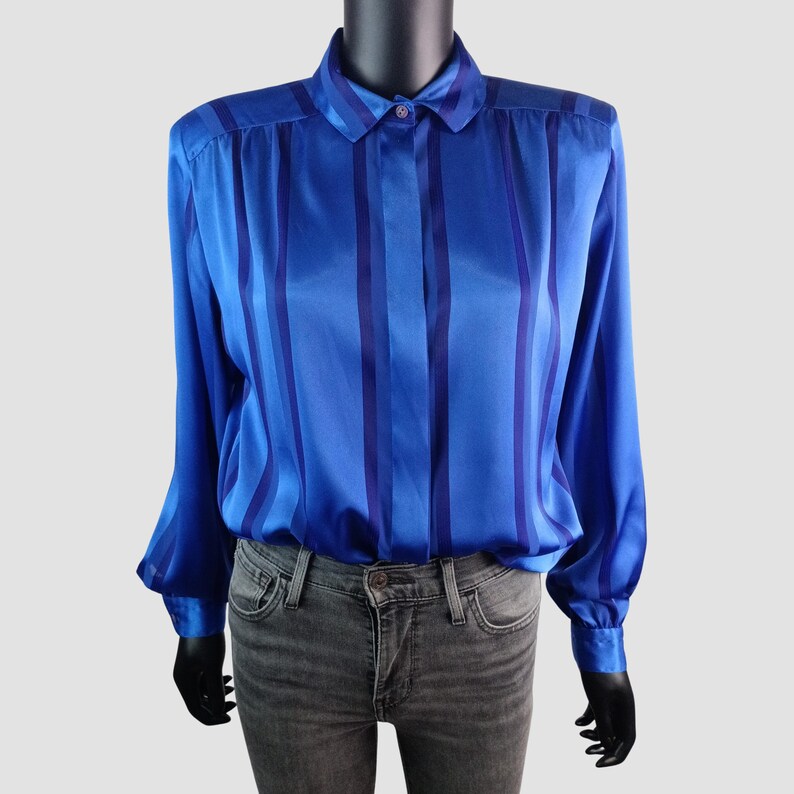 May include: A vibrant blue blouse with vertical stripes in a darker shade of blue. The long-sleeved shirt features a classic collar and buttoned cuffs. The blouse is made of a shiny, silky material.