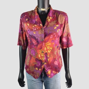 May include: A button-down blouse with a vibrant floral pattern in shades of red, orange, purple, and yellow. The short-sleeved shirt has a collared neckline and is displayed on a mannequin. The blouse is paired with blue jeans.