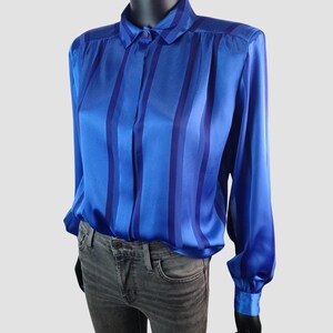 May include: A blue satin blouse with vertical stripes in varying shades of blue. The long-sleeved shirt has a button-down front, a classic collar, and buttoned cuffs. The blouse is displayed on a mannequin.