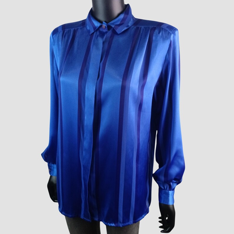 May include: A long-sleeved blue blouse with vertical stripes in varying shades of blue. The blouse has a collared neckline, buttoned cuffs, and a silky, shiny texture. The garment is displayed on a mannequin.