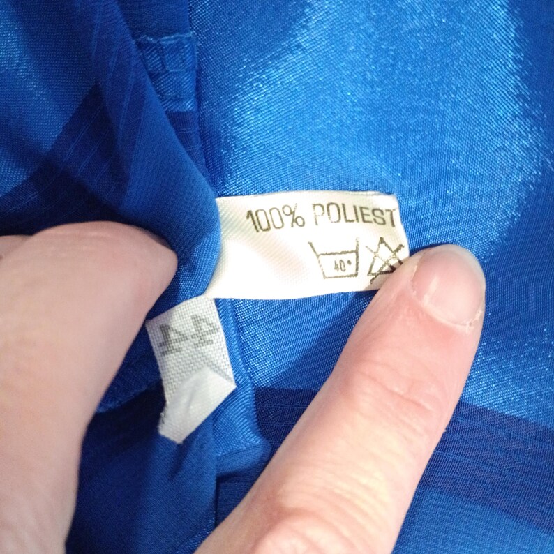 May include: Close-up of a white clothing tag with the text "100% POLIEST" and washing instructions. The tag is attached to a vibrant blue fabric. The tag also has a symbol indicating a 40-degree wash.