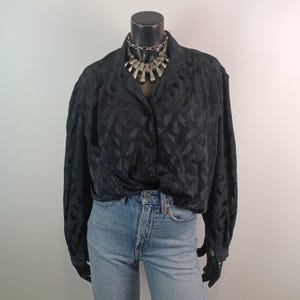 以下が含まれることがあります： 黒いボタンダウンブラウスは、繊細な筆使いのパターンで、明るい色のデニムジーンズと組み合わせています。シルバーのステートメントネックレスが華やかさを添えています。ブラウスは長袖で、マネキンに着せています。