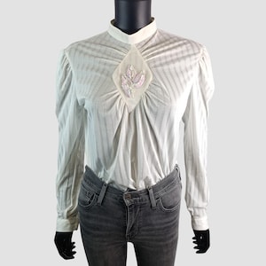 Può includere: Una camicetta bianca vintage con collo alto e maniche lunghe. La camicetta presenta strisce verticali e un abbellimento a forma di diamante sul petto con paillettes rosa e bianche. Le maniche hanno una leggera sbuffatura sulle spalle.