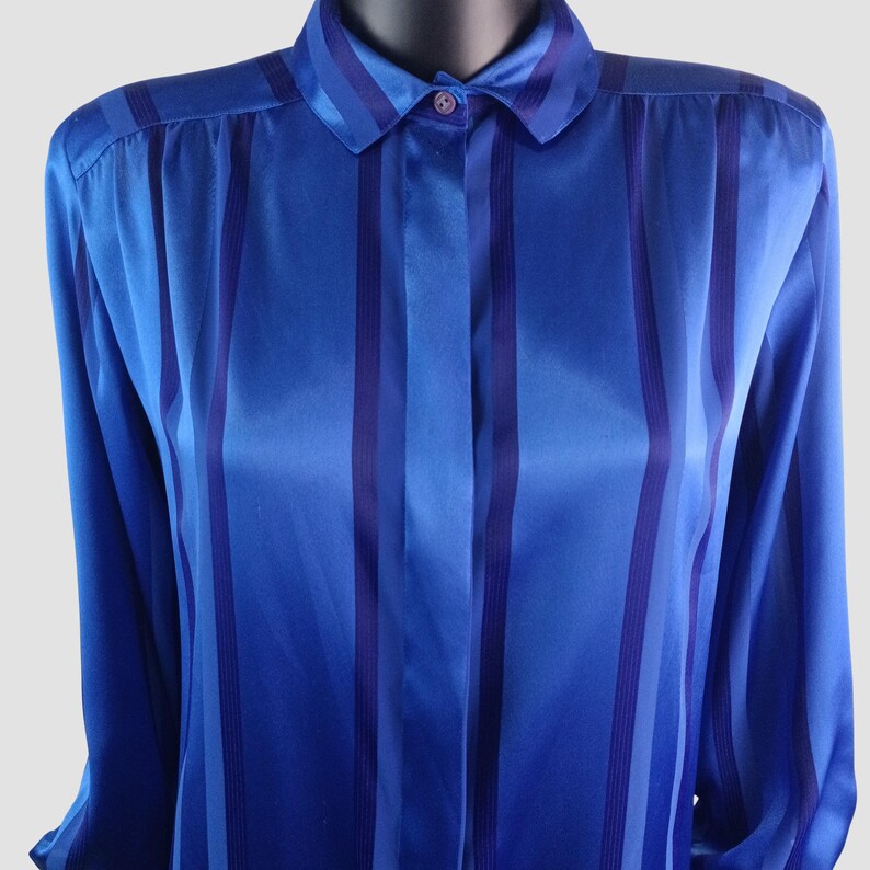 May include: A blue button-down shirt with vertical stripes in varying shades of blue. The long-sleeved shirt has a classic collar and is made of a shiny, silky material. The shirt is displayed on a mannequin.