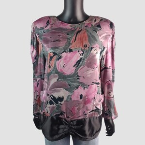 Peut inclure: Un chemisier à manches longues avec un motif floral dans les tons de rose, gris et orange. Le chemisier a un bas en satin noir avec un détail froncé. Le haut est présenté sur un mannequin noir.