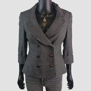 Puede incluir: Un blazer gris de doble botonadura con un sutil estampado de cuadros. El blazer presenta botones marrones oscuros, un cuello con solapas y mangas tres cuartos. Se ve un collar plateado con un colgante.