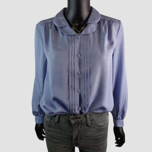 Puede incluir: Una blusa azul claro con cuello redondo y mangas largas. La parte delantera de la blusa presenta pliegues verticales y se abrocha con botones. La blusa se combina con vaqueros grises. Se ve un collar plateado.