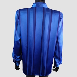 May include: A long-sleeved blue shirt with vertical stripes in varying shades of blue. The shirt has a collared neckline and buttoned cuffs. The fabric has a satin-like sheen, giving it a smooth and elegant appearance. The shirt is displayed on a mannequin.