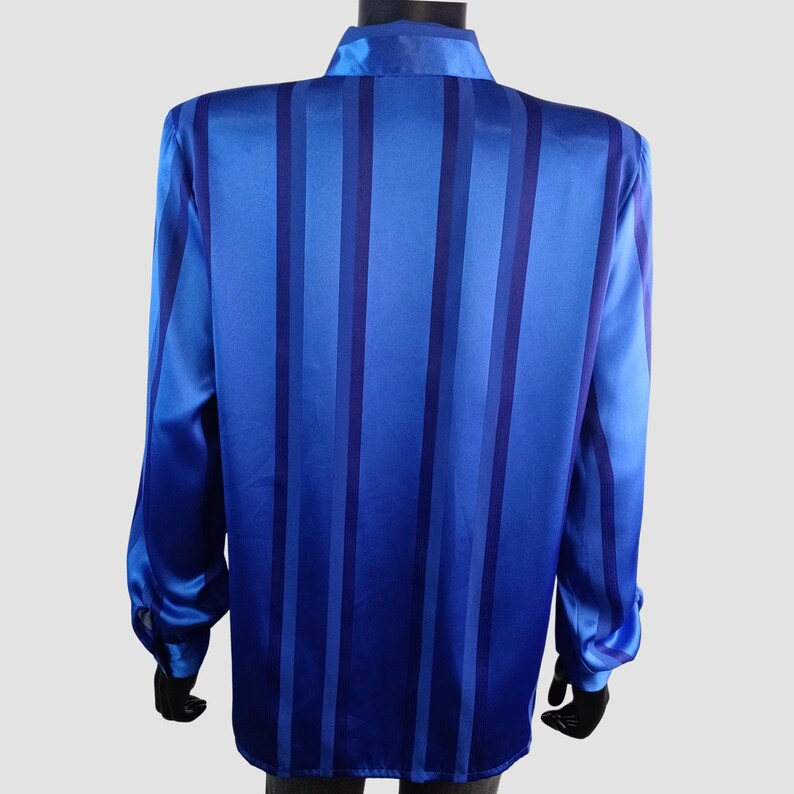 May include: A long-sleeved blue shirt with vertical stripes in varying shades of blue. The shirt has a collared neckline and buttoned cuffs. The fabric has a satin-like sheen, giving it a smooth and elegant appearance. The shirt is displayed on a mannequin.