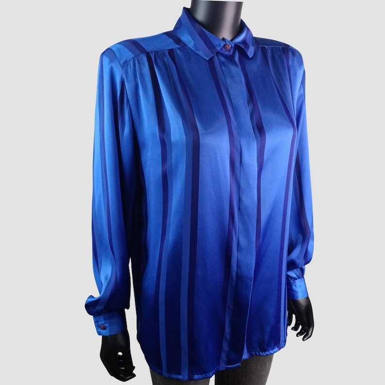 May include: A blue satin button-down shirt with vertical stripes in varying shades of blue. The long-sleeved shirt has a collared neckline and buttoned cuffs. The shirt is displayed on a mannequin.