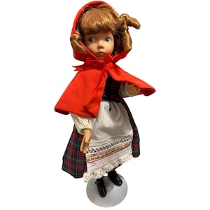 Puede incluir: Muñeca vintage vestida con una capa con capucha roja, blusa blanca y falda a cuadros. La muñeca tiene cabello castaño rizado y está de pie sobre una esfera blanca. La falda tiene un delantal blanco con ribete bordado y encaje.