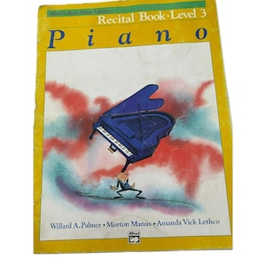 Op de afbeelding: Een gele boekomslag met de titel "Recital Book - Level 3 Piano" en een cartoonillustratie van een persoon die een blauwe piano boven zijn hoofd houdt.