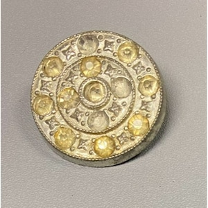 Puede incluir: Un botón plateado con un diseño circular que presenta un círculo central elevado rodeado de círculos más pequeños y un borde de estrellas. El botón está decorado con diamantes de imitación transparentes.