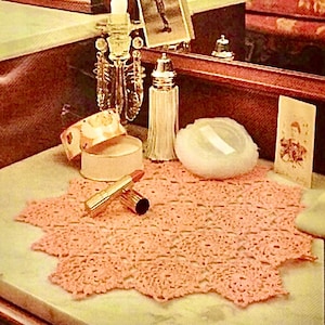 Könnte beinhalten: Ein Stillleben mit einem rosa gehäkelten Deckchen, Lippenstift, einem Puderquaste und einer kleinen runden Schachtel. Eine Kerze in einem Kristallhalter, ein Salzstreuer und gerahmte Fotos sind ebenfalls vorhanden. Die Gegenstände sind auf einer Marmoroberfläche angeordnet.