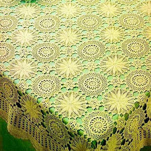 May include: A crocheted tablecloth with a delicate floral pattern in a cream colour. The tablecloth is laid on a green table and features a repeating pattern of circular motifs.