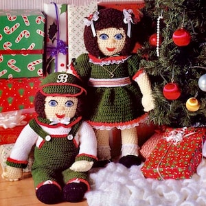 May include: Two handmade crochet dolls, one seated and one standing, are featured in this image. The seated doll wears green overalls and a cap with a "B". The standing doll is dressed in a green dress. Both dolls have blue eyes and are near a Christmas tree.